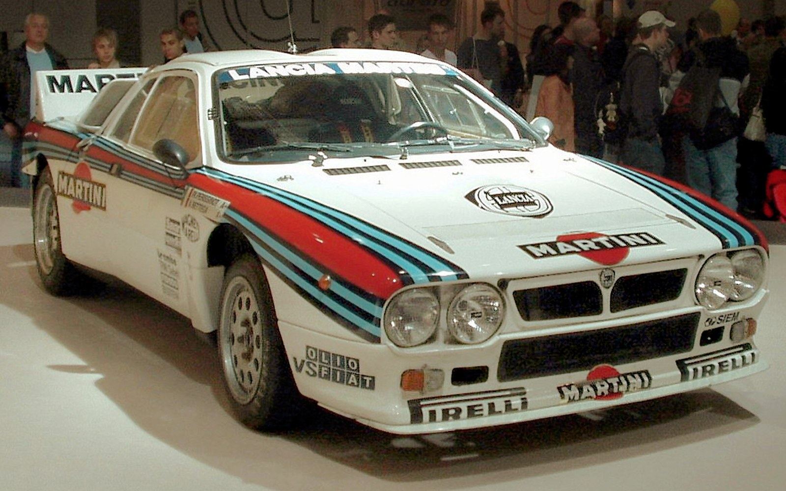 A Group B rally car of the mid-1980s at speed on a gravel stage, boxed arches and large rear wing, dust behind